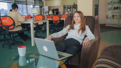 Poster - Young woman working at the open space office. She sitting at the chair near coffee table. Female typing on laptop. Adult lady with long red hair finish her project and leans back in a chair. She smile