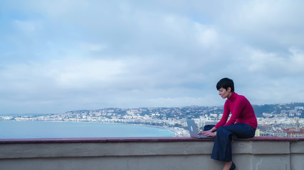 Poster - Female sitting in front of sea and slouch. Caucasian model wearing in jeans and red pullover using laptop outdoor. On the background seascape with sea beach and seafront in the marine city.