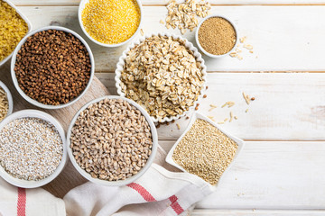 Selection of whole grains in white bowls - rice, oats, buckwheat, bulgur, porridge, barley, quinoa, amaranth, on white wood background