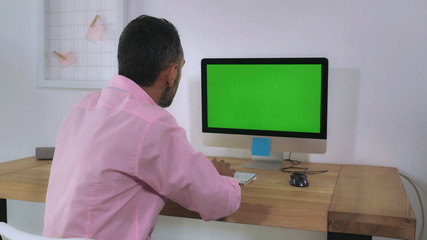 Leinwandbilder - Back view focused businessman wearing casual shirt sitting at the working place works on computer with green display. Man entering data typing on keyboard in home office looking on pc