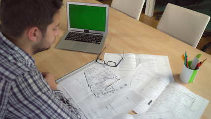 Aufkleber - Man designer working with blueprint at the working place. On the desk laptop with green screen. Male holding pen and checking and drawing