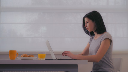 Poster - Young woman typing on laptop in the morning. Female with black hair wearing in casual t-shirt sitting at the table with breakfast in front of big window.