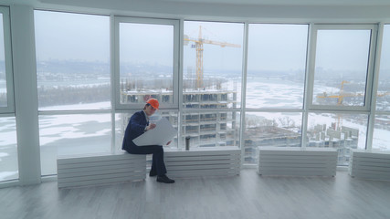 Poster - happy building engineer sitting in the modern office looking on the blue print documents with architectural plan. handsome man wearing in elegant suit and hard hat. At the construction site workers