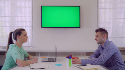 Poster - Coworkers sitting at the white desk talking discussing project. Two adult people wearing in stylish colored clothes. On the wall display with green screen.