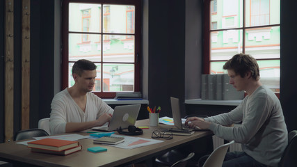 Aufkleber - Two young businessmen working in the modern contemporary office with big windows. Group of people works with computer sitting at the wooden desk with paper documents. Happy startup team.