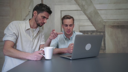 Poster - Friends drink coffee and tea consider the pros and cons of project. Mixed race man smiling. Caucasian man gesturing and point on the laptop monitor.