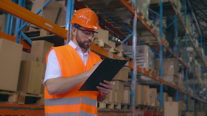 Poster - Handsome young employee working at warehouse. Worker wearing high visibility clothing and a hard hat checking and count up goods or boxes for delivery. Man writing some notes.