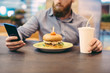 © ruslanshug - young bearded man in shirt sitting in cafe with burger and paper cup holding smartphone online