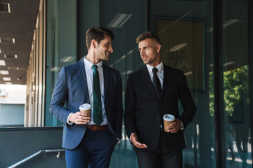 Sticker - Image of caucasian businessmen partners talking and drinking takeaway coffee while standing outside office center