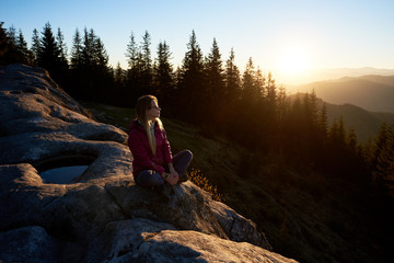 Houng woman traveller sitting on big rock on the top of mountain in the morning. Active female tourist enjoying amazing sunrise. On background forest, rising sun and blue sky