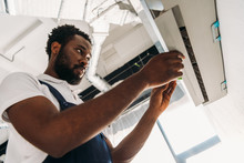 Concentrated African American Repairman Standing On Ladder And Fixing Air Conditioner