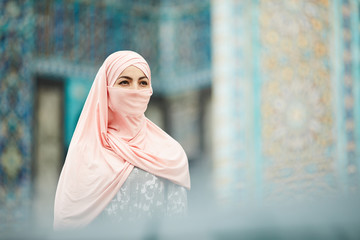 Wall Mural - Introspective young Muslim woman in pink hijab standing against mosque with bright design and looking into distance