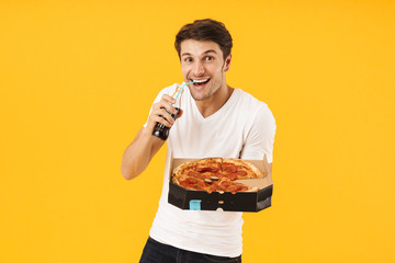 Poster - Cheerful happy young man in casual white t-shirt eat pizza drinking soda isolated over yellow background.