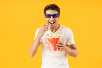 Young man in casual white t-shirt eat popcorn wearing 3d glasses watch film isolated over yellow background.