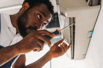 serious african american repairman fixing air conditioner with screwdriver