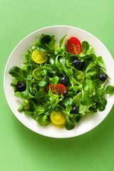 fresh salad of arugula and valerian leaves with colored tomatoes and blueberries in a plate on a green background.