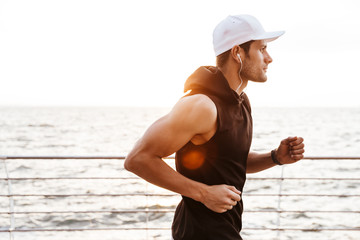 Canvas Print - Photo of athletic man in white cap listening to music with earphones while jog on wooden pier at seaside