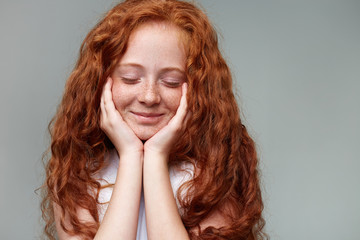 Wall Mural - Close up of nice little dreamily girl with ginger hair and freckles, stands over gray background with closed eyes and touches cheeks.