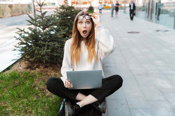 Poster - Shocked young business woman posing sitting outdoors near business center wearing eyeglasses using laptop computer.