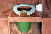 Toilet Seat Of A Country Wooden Outdoor Toilet With A Metal Bucket As A Waste Tank And A Roll Of A Paper Close Up