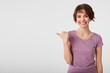 © timtimphoto - Cute cheerful short haired young woman in blank t-shirt, lloks happy and broadly smiles, wonts to draw you attention and points fingers to the copy space on the left side over white wall.