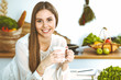 © rogerphoto - Young happy woman is holding white cup and looking at the camera while sitting at wooden table in the kitchen among green vegetables