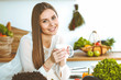 © rogerphoto - Young happy woman is holding white cup and looking at the camera while sitting at wooden table in the kitchen among green vegetables