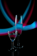 A two wine glass isolated on a black background with red, white, pink and orange neon light painting streaks of light behind them. Light wave, play of color, drawing with light.