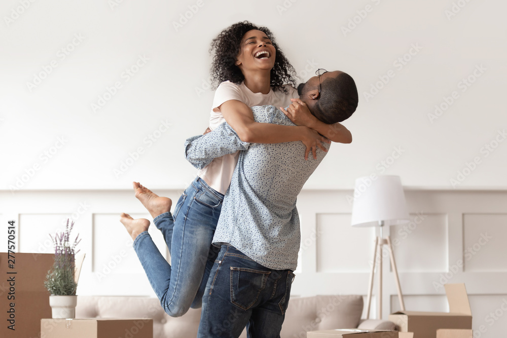 African husband lifting happy wife celebrating moving day with boxes - obrazy, fototapety, plakaty 