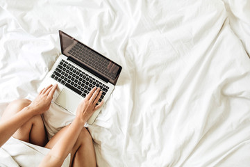 Wall Mural - Working at home concept. Girl is working on her laptop in bed with white linens. Flat lay, top view minimalistic workspace background. Relaxing working process.