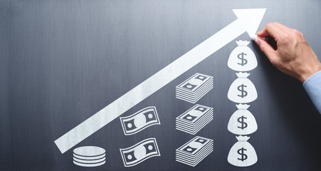 Saving and growing money concept. Businessman drawing money and arrow on chalkboard.