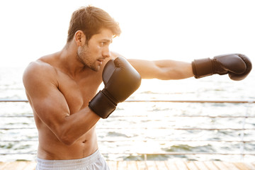 Poster - Photo of masculine half-naked man working out in black boxing gloves on wooden pier at seaside