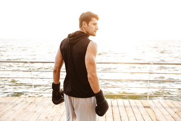 Wall Mural - Photo of serious man in tracksuit working out in black boxing gloves on wooden pier at seaside