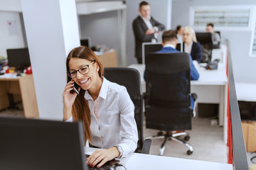 Wall Mural - Gorgeous Caucasian businesswoman in formal wear using computer and talking on the phone. In background her coworkers working.