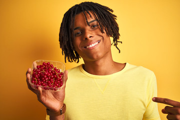 Afro american man with dreadlocks holding red currants over isolated yellow background with surprise face pointing finger to himself