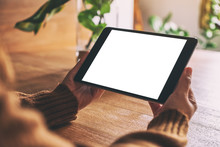 Mockup Image Of Hands Holding And Using Black Tablet Pc With Blank White Desktop Screen On Wooden Table