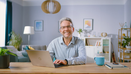 handsome middle aged man uses laptop computer and smiles on camera. he is in the cozy and stylish li