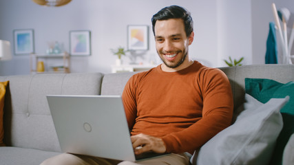 Wall Mural - Cheerful Young Man at Home Sitting on a Sofa Holds Laptop on His Lap, Browses Through the Internet, Social Networks, Does e-Shopping.