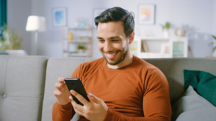 Happy Young Man Uses Smartphone while Sitting on a Sofa at Home. Man Browses Through Internet, Watches Videos and Uses Social Networks at Home.