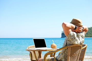 Fit travel blogger sitting at beach bar writing article on white laptop. Freelance remote work concept. Self employed man coding, wearing typical tourist shirt and hat. Copy space, sea view background