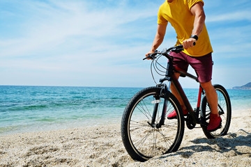 Young man with fit body riding the mtb mountain bike on sandy beach with beautiful azure water sea view. Muscle male wearing bright yellow t-shirt cycling on ocean shore. Close up, copy space.