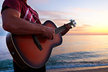 Young Man Wearing Purple Tie Dye T-shirt Playing Dreadnought Parlor Acoustic Guitar On Beach At Beautiful Sunset Time. Fit Guitarist W/ Sunburst Instrument By The Sea. Background, Copy Space, Close Up