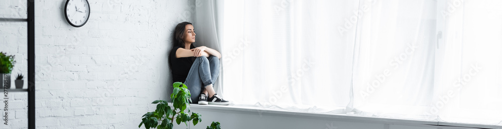 panoramic shot of depressed young woman sitting on window sill with crossed arms and looking away - obrazy, fototapety, plakaty 