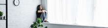 Panoramic Shot Of Depressed Young Woman Sitting On Window Sill With Crossed Arms And Looking Away