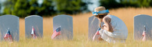 Panoramic Shot Of Kid In Dress And Straw Hat Sitting Near Headstone With American Flag In Graveyard