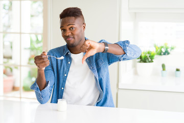 Sticker - African american man eating healthy natural yogurt with a spoon with angry face, negative sign showing dislike with thumbs down, rejection concept