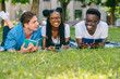 © Iryna - Multi-ethnic group of students friends lying on grass talking laughing reading books while doing homework at park in sunny meadow. Education and Friendship Concept. Black woman laughing at funny joke.