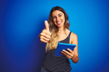 Poster - Young beautiful woman using touchpad tablet over blue isolated background happy with big smile doing ok sign, thumb up with fingers, excellent sign