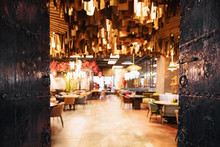 Blurred Image Of The Interior Of A Large Beautiful Restaurant With Bright Lighting. The Waitress Comes To The Working Bar And The Bartender, Gives Visitors A Menu And Goes For A Drink. Defocused Staff