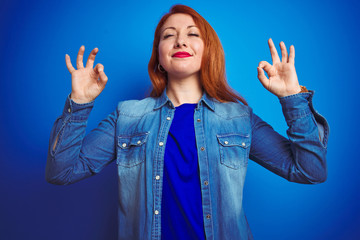 Sticker - Young beautiful redhead woman wearing denim shirt standing over blue isolated background relaxed and smiling with eyes closed doing meditation gesture with fingers. Yoga concept.